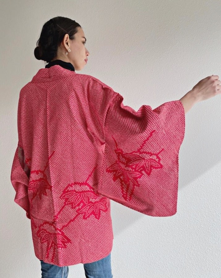 A woman wearing a black turtleneck and jeans is seen from behind wearing a red color in the product Bamboo Leaves Shibori Haori Kimono Jacket by KIMONO ZEN brand, shades of red.