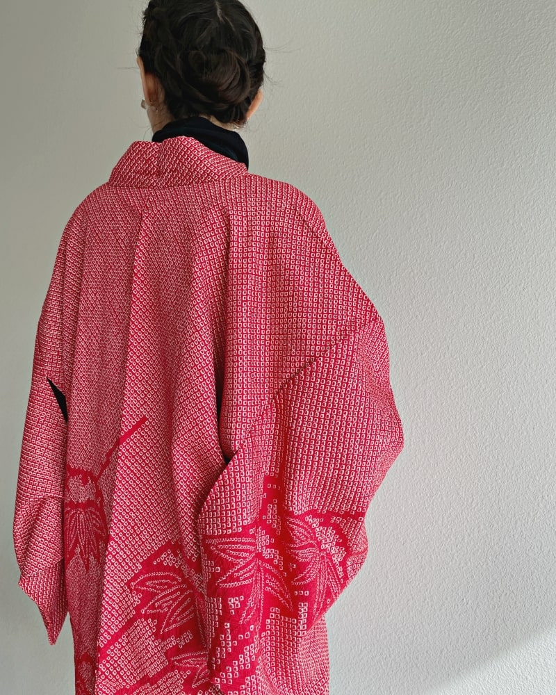 A woman wearing a black turtleneck and jeans is seen from behind wearing a red color in the product Bamboo Leaves Shibori Haori Kimono Jacket by KIMONO ZEN brand, shades of red.