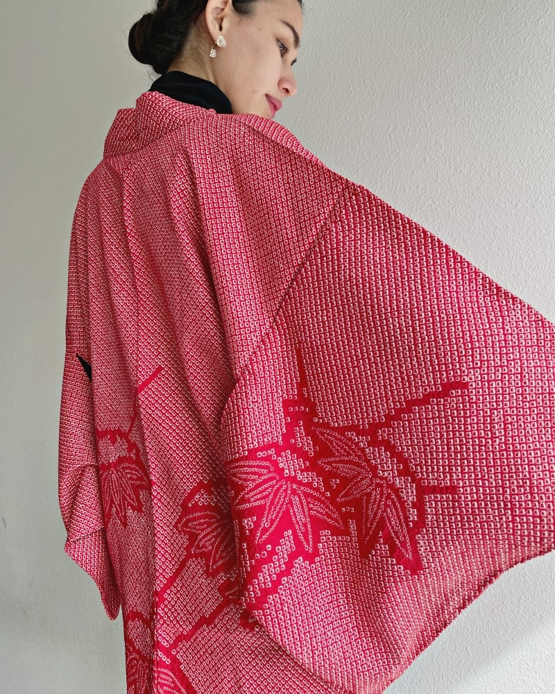 A woman wearing a black turtleneck and jeans is seen from behind wearing a red color in the product Bamboo Leaves Shibori Haori Kimono Jacket by KIMONO ZEN brand, shades of red.