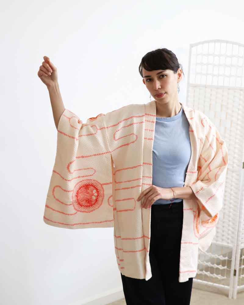 A woman wearing a Powder Pink kimono, facing forward and rising right hand.