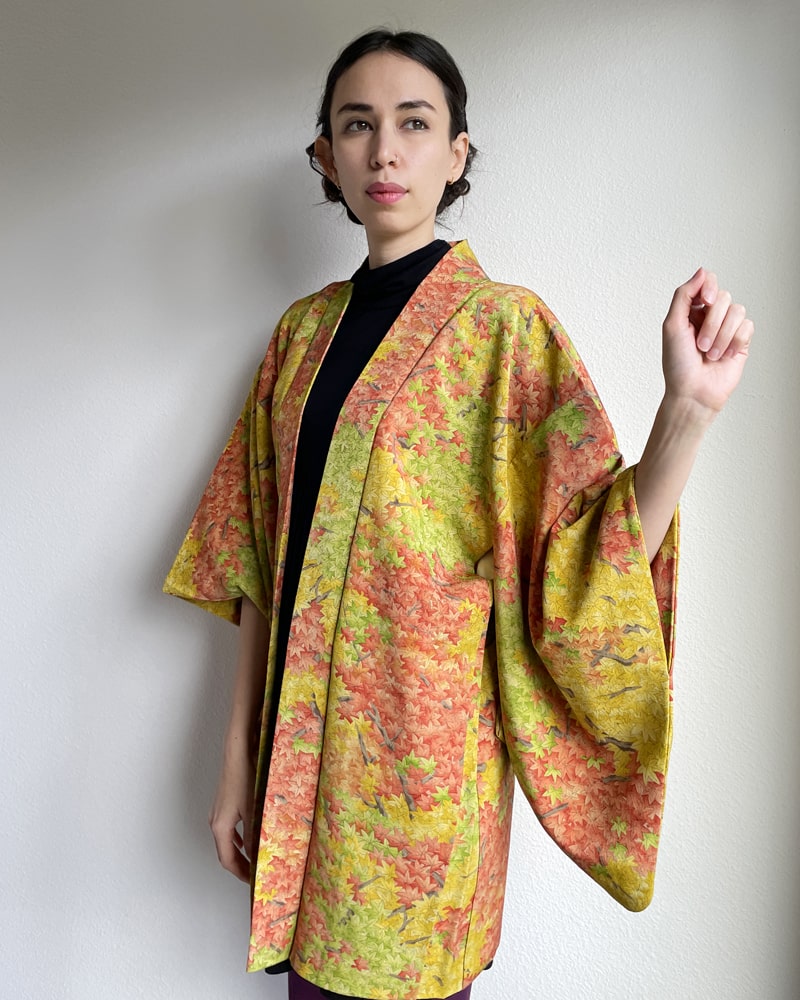 Front oblique side view of a woman wearing a KIMONO ZEN brand Autumn Leaves Forest Haori Kimono Jacket, which is a mixture of yellow and green in hue with autumn color lettering throughout.