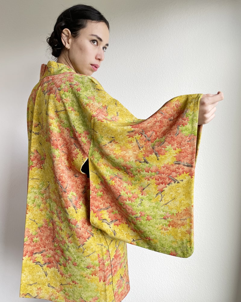Side view of a woman wearing a KIMONO ZEN brand Autumn Leaves Forest Haori Kimono Jacket, which is a mixture of yellow and green in hue with autumn color lettering throughout.
