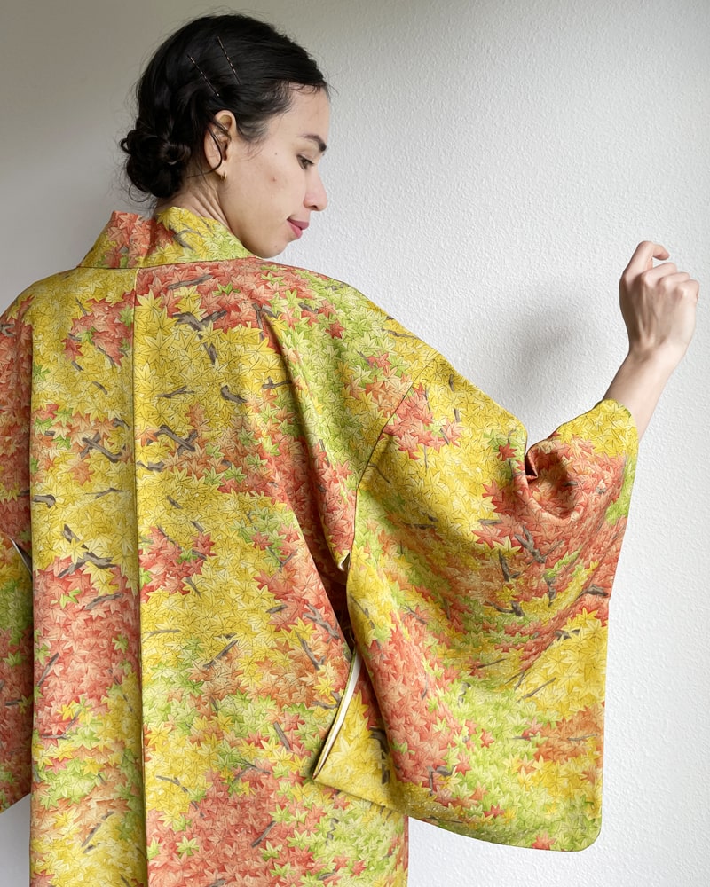 A woman wearing a KIMONO ZEN brand Autumn Leaves Forest Haori Kimono Jacket, a mixture of yellow and green in hue with autumn color lettering throughout, seen from behind.