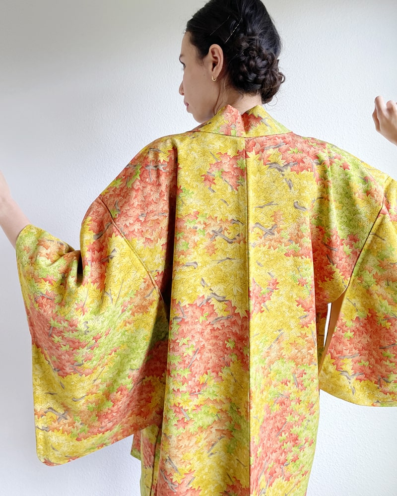 A woman wearing a KIMONO ZEN brand Autumn Leaves Forest Haori Kimono Jacket, a mixture of yellow and green in hue with autumn color lettering throughout, seen from behind.