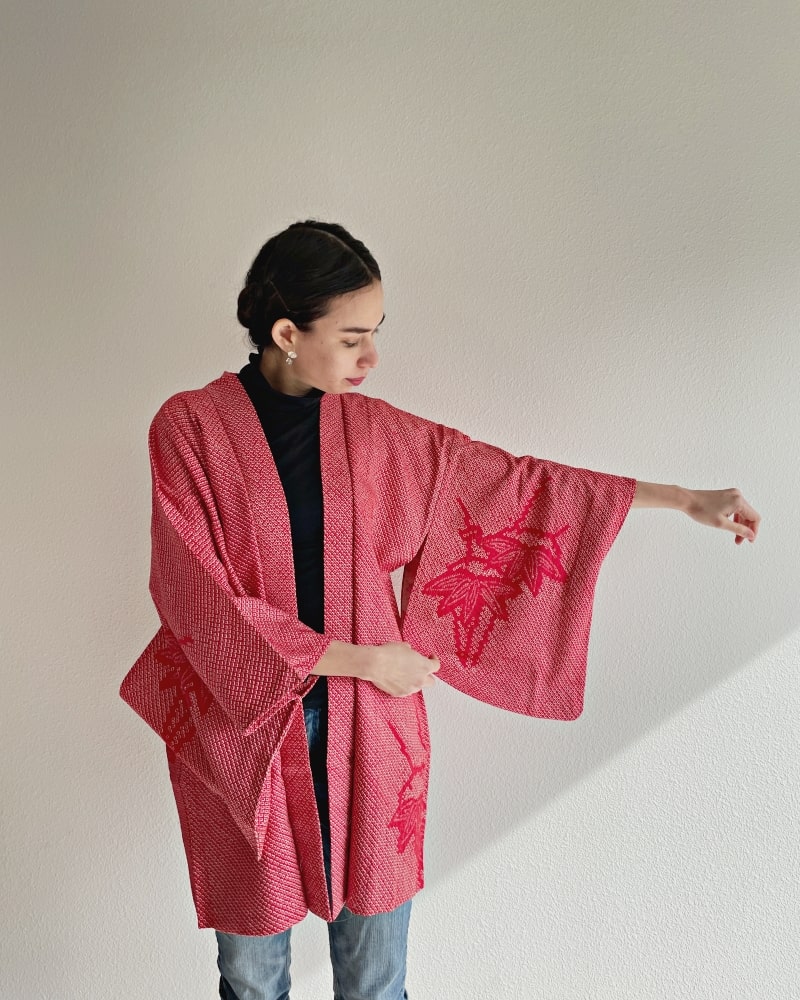 Front view of a woman wearing a black turtleneck and jeans wearing a red color in the product Bamboo Leaves Shibori Haori Kimono Jacket by KIMONO ZEN brand, shades of red.