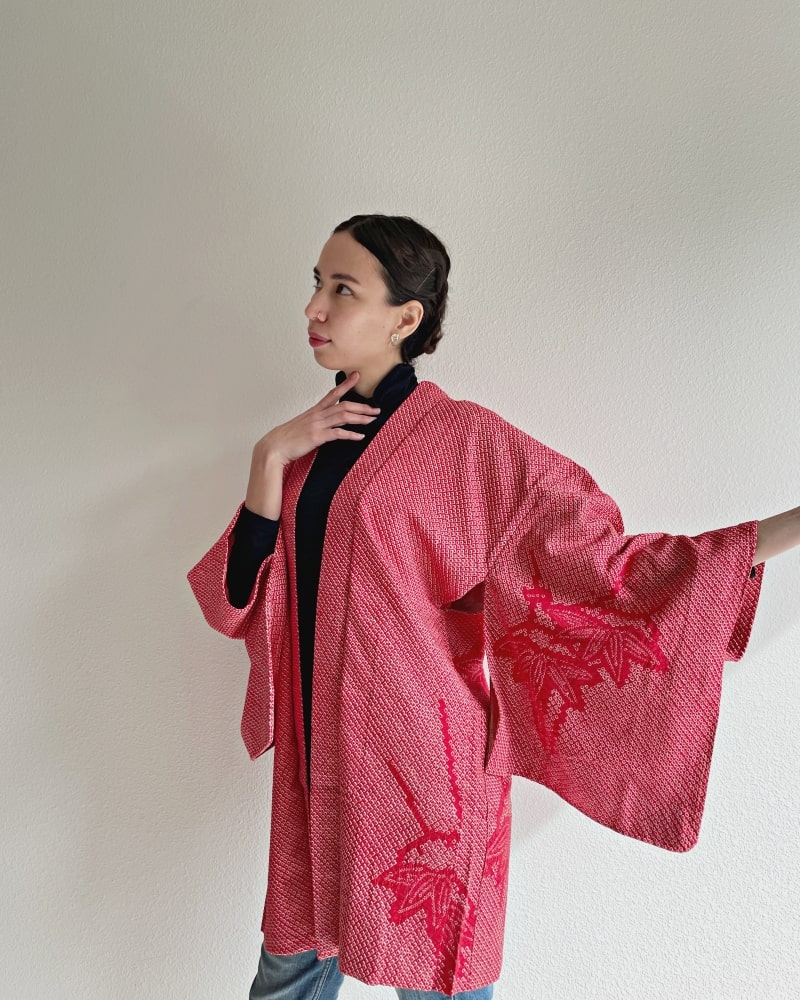 A woman wearing a black turtleneck and jeans wearing a red color in the Bamboo Leaves Shibori Haori Kimono Jacket product of the KIMONO ZEN brand.