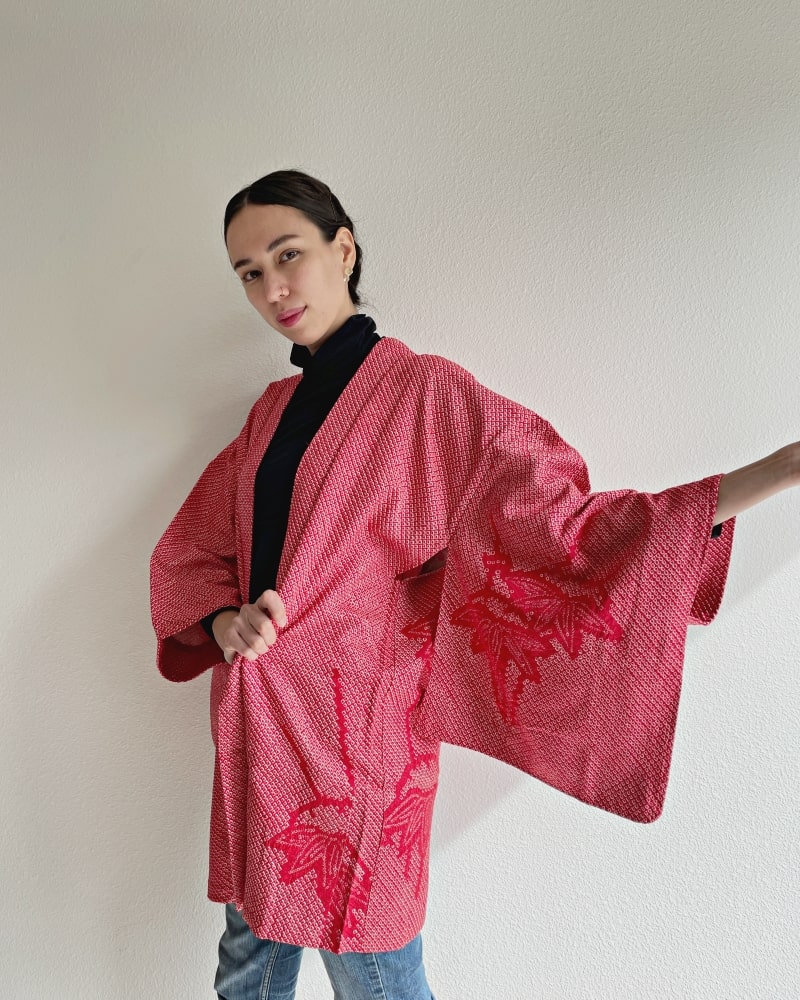 A woman wearing a black turtleneck and jeans wearing a red color in the Bamboo Leaves Shibori Haori Kimono Jacket product of the KIMONO ZEN brand.