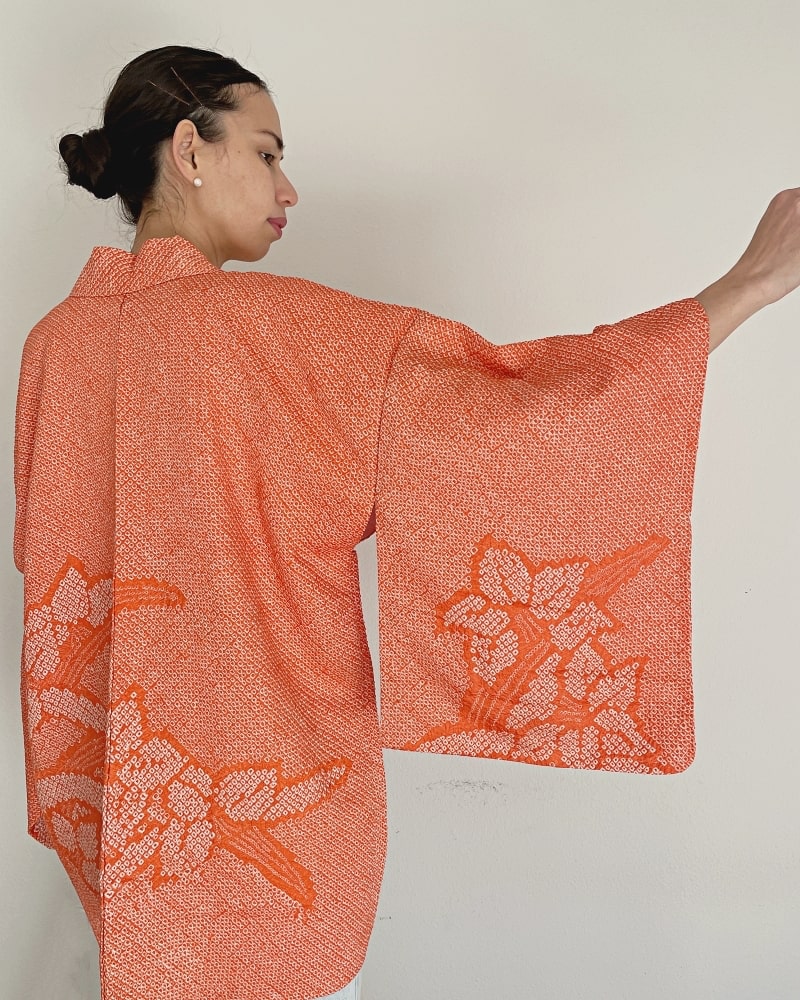 A woman's upper body is seen from behind wearing a beautiful autumn-colored shibori fabric with a white cut and a pair of jeans, in a product called Beautiful Narcissus Haori Kimono Jacket by Kimono zen brand.