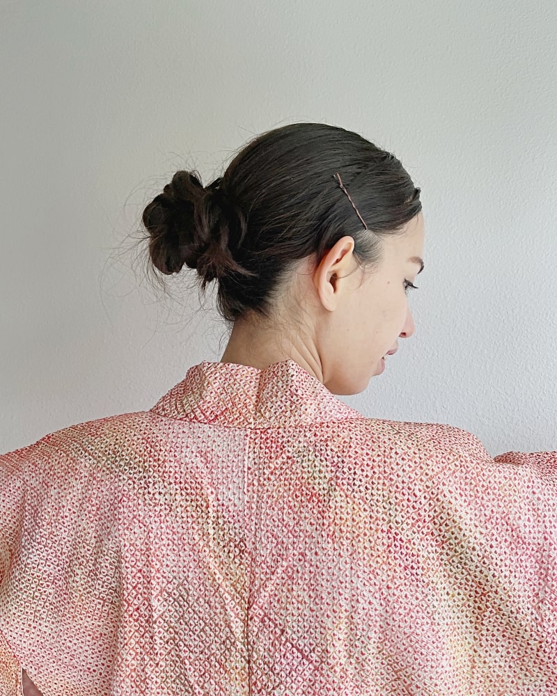 A woman wearing a pinkish-colored shibori fabric with a plum pattern on the back collar of a woman wearing a white T-shirt and jeans with the Bell Flower Love Shibori Haori Kimono Jacket from the Kimono zen brand.