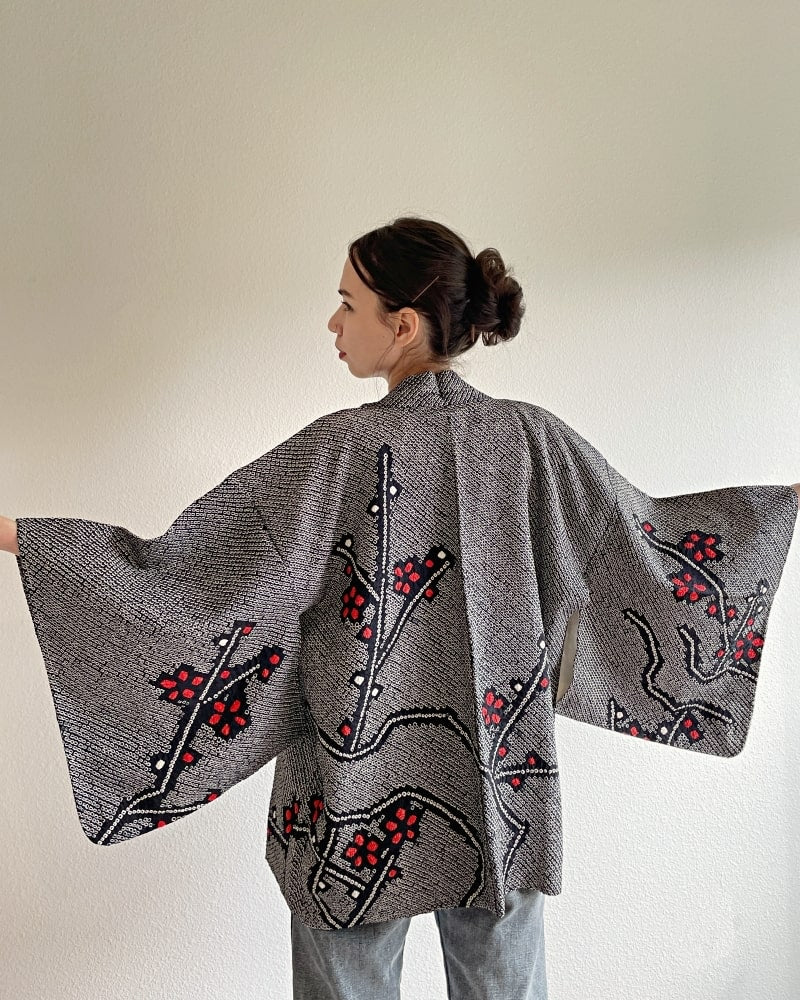 A woman wearing a Kimono zen brand Black Cherry Blossom Shibori Haori Kimono Jacket, a black shibori fabric jacket with a bright red plum pattern, with a white cut and jeans.