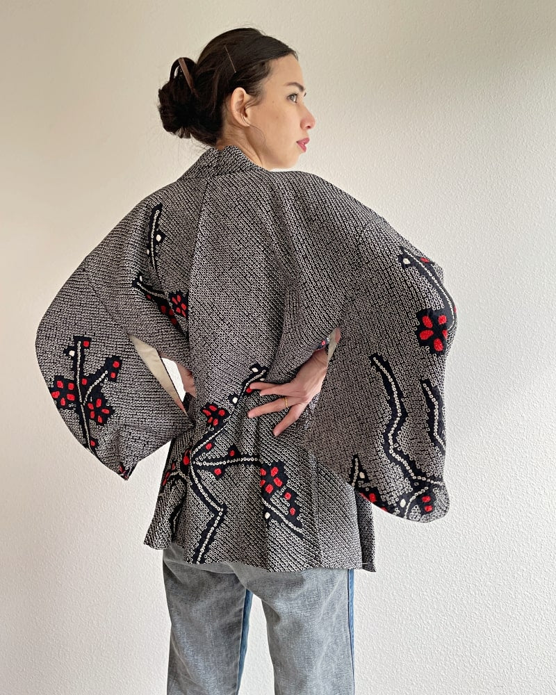 A woman wearing a Kimono zen brand Black Cherry Blossom Shibori Haori Kimono Jacket, a black shibori fabric jacket with a bright red plum pattern, with a white cut and jeans.