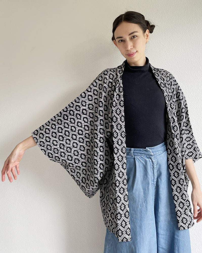 Black Shibori Haori Kimono Jacket of the Kimono zen brand, black in color with diamond-shaped pattern all over, worn by a woman with a black turtleneck and jeans on her upper body