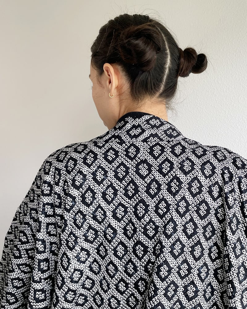 Back view of a woman wearing a Kimono zen brand Black Shibori Haori Kimono Jacket, black in color with a diamond-shaped pattern all over it, with a black turtleneck and jeans.
